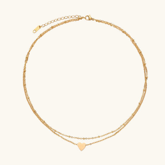 Two-layer gold-plated stainless steel necklace with small heart pendant displayed on beige background