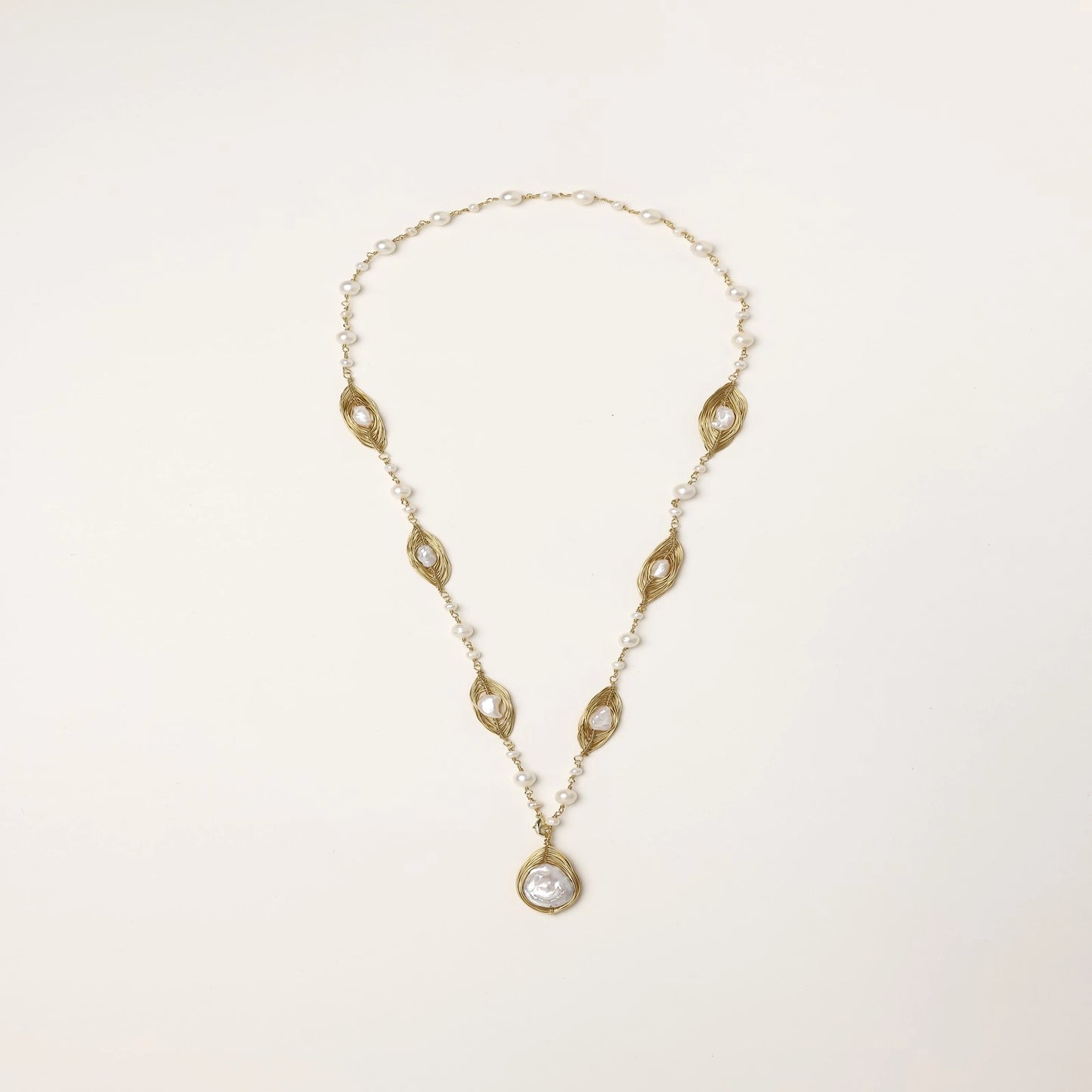 Long gold-plated necklace with eye-shaped elements, freshwater pearls, and baroque pearl pendant on neutral background.