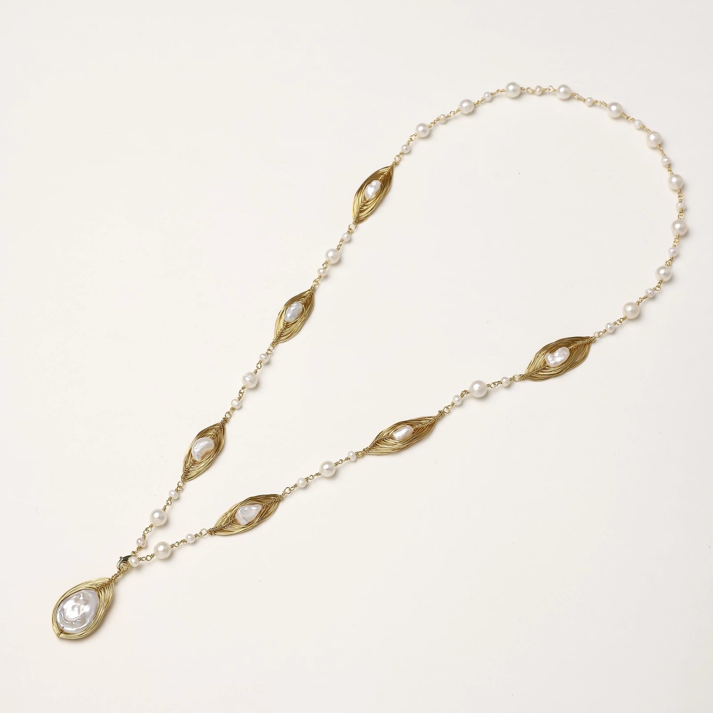 Long gold-plated necklace with eye-shaped elements, freshwater pearls, and baroque pearl pendant on neutral background.