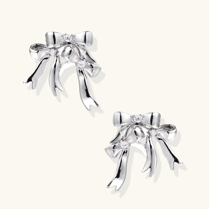 Pair of 18K gold plated brass bow-shaped earrings with flowing ribbon tails and zirconia crystal accents on white background.
