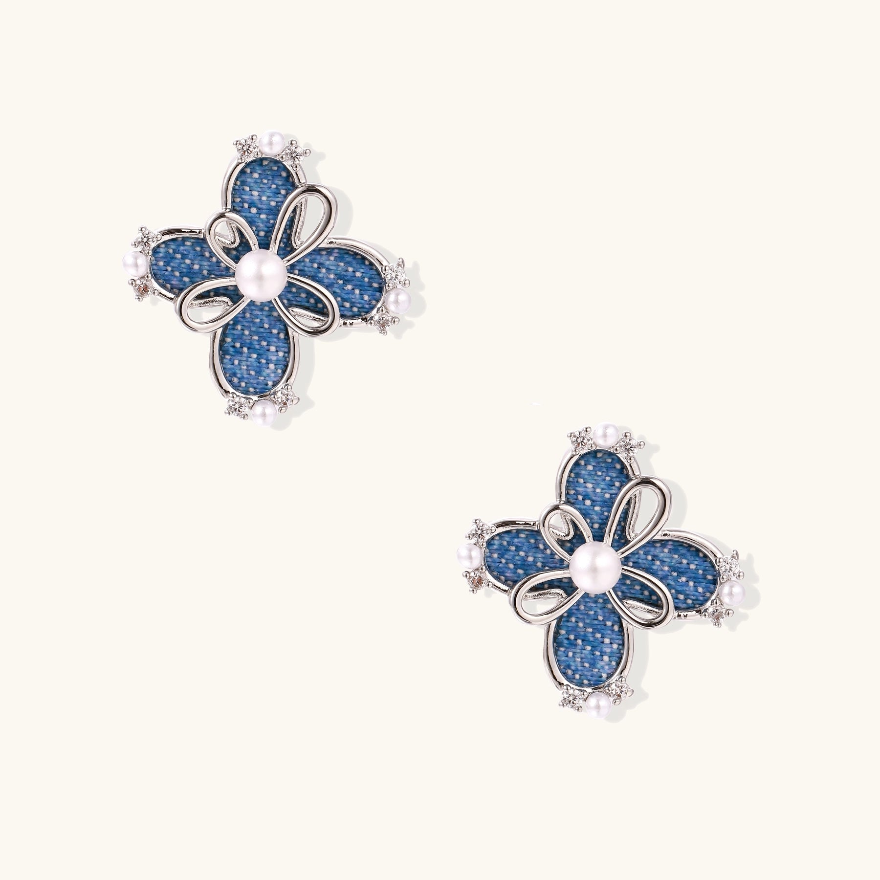 Blue denim flower stud earrings with silver framework, white pearl centers, and crystal accents on cream background.