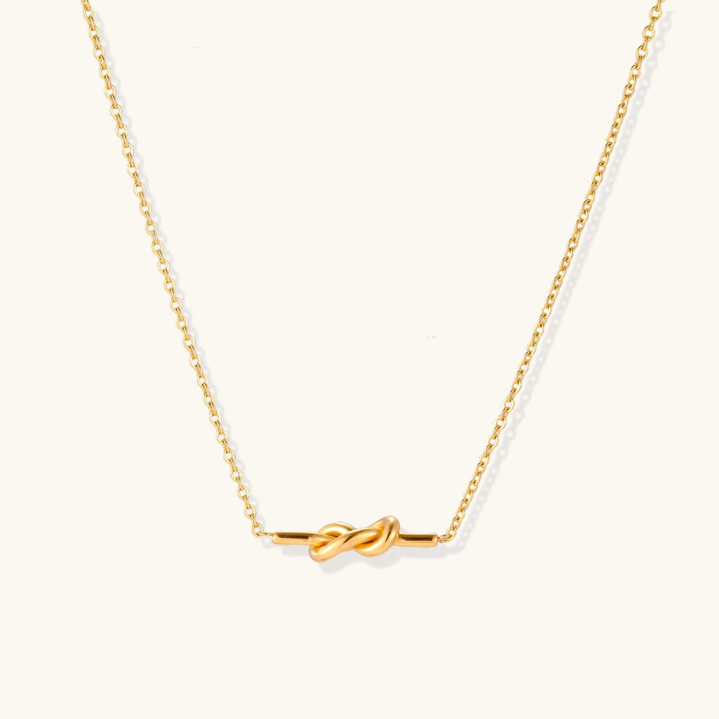 Dainty 18K gold plated stainless steel chain necklace with minimalist knot pendant on cream background.