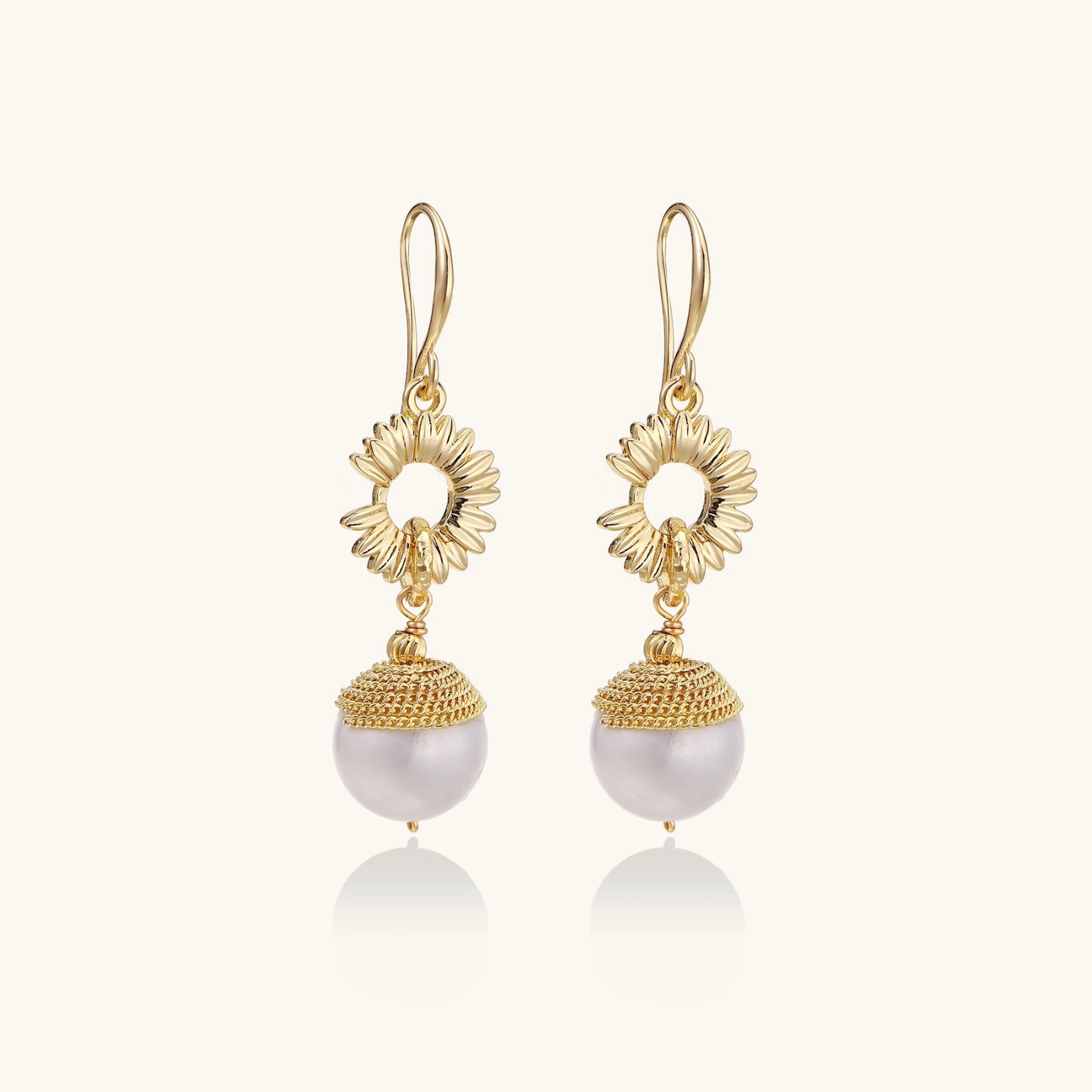 Gold sunburst charm earrings with baroque pearl drops on French hooks against cream background