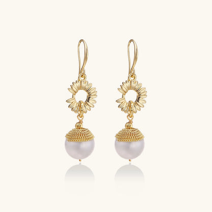 Gold sunburst charm earrings with baroque pearl drops on French hooks against cream background