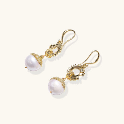 Gold sunburst charm earrings with baroque pearl drops on French hooks against cream background