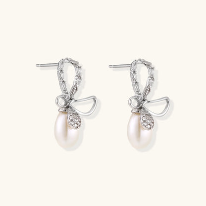 Silver bow-shaped stud earrings with baguette crystals and dangling white freshwater pearls on cream background