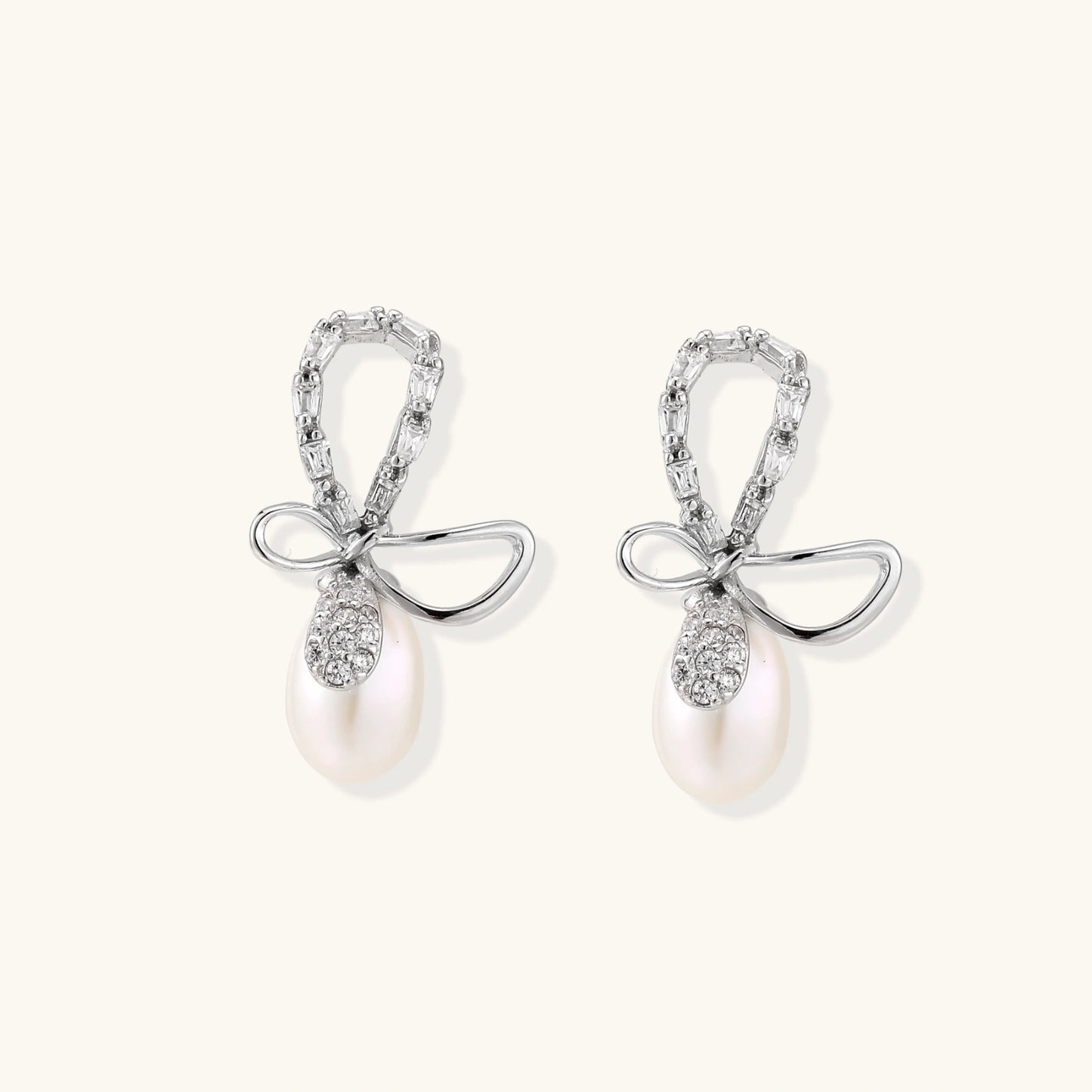 Silver bow-shaped stud earrings with baguette crystals and dangling white freshwater pearls on cream background