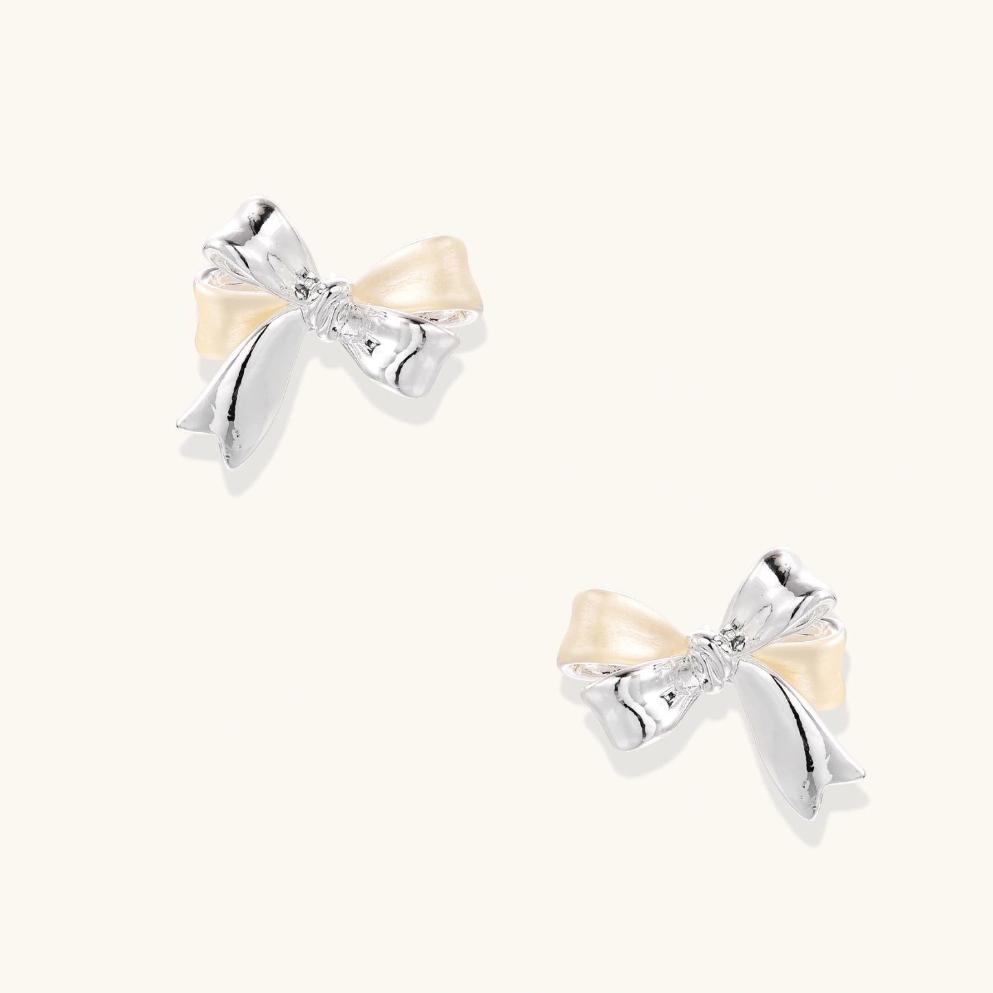 Dimensional bow stud earrings in white gold plated brass with silver and cream two-tone finish on neutral background