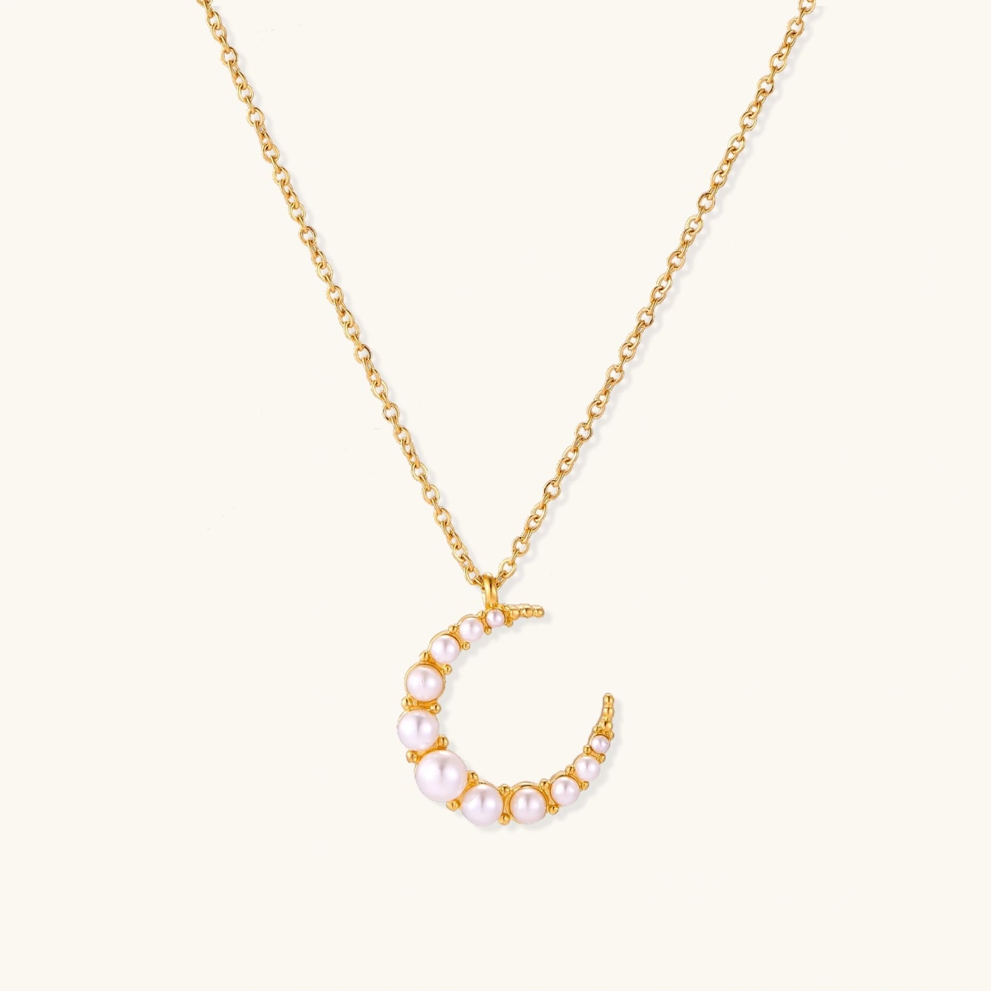 Gold crescent moon pendant with graduated white pearls on delicate chain against cream background