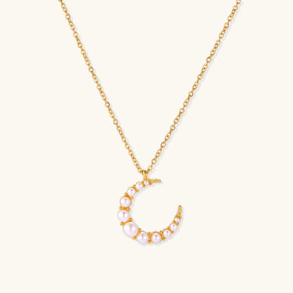 Gold crescent moon pendant with graduated white pearls on delicate chain against cream background