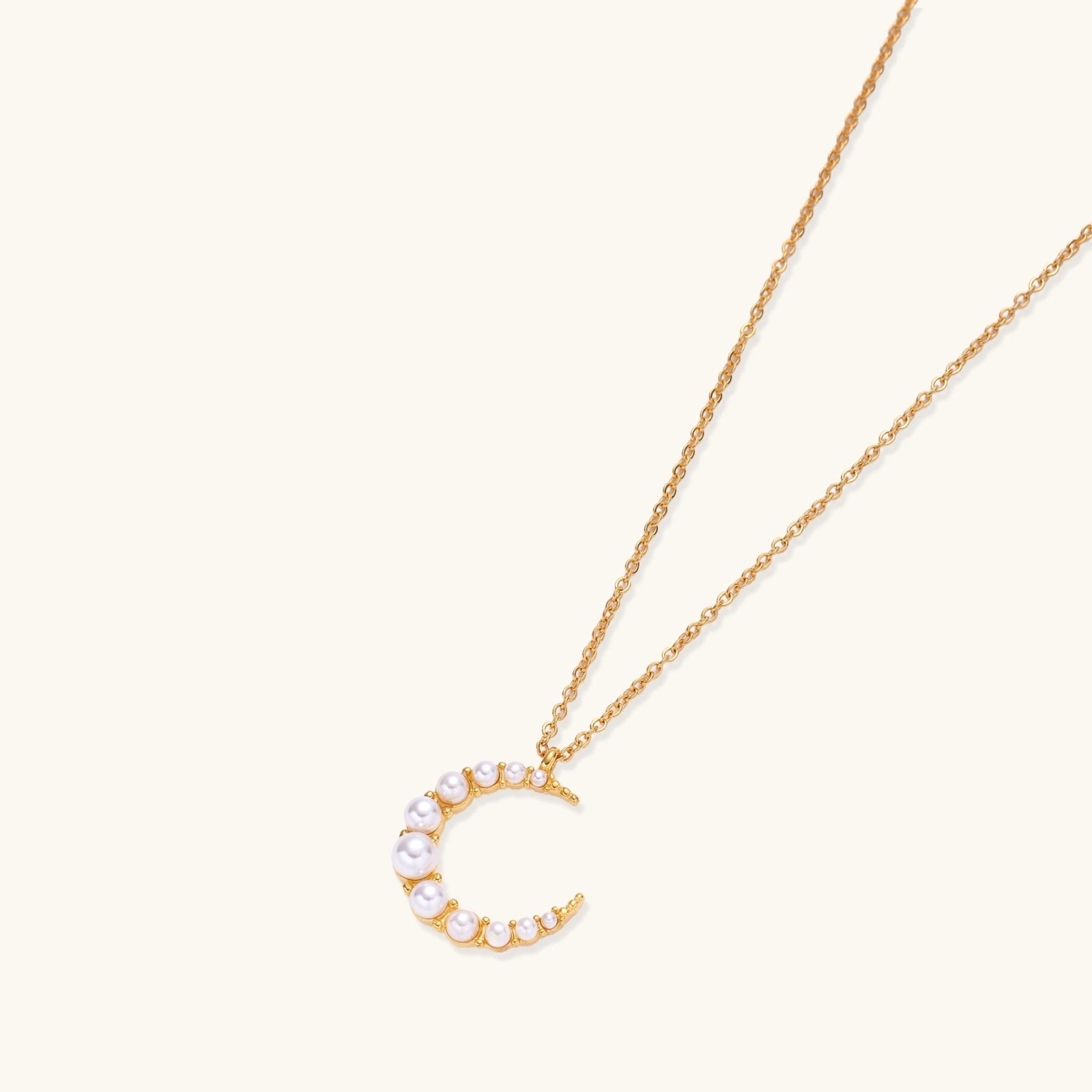 Gold crescent moon pendant with graduated white pearls on delicate chain against cream background