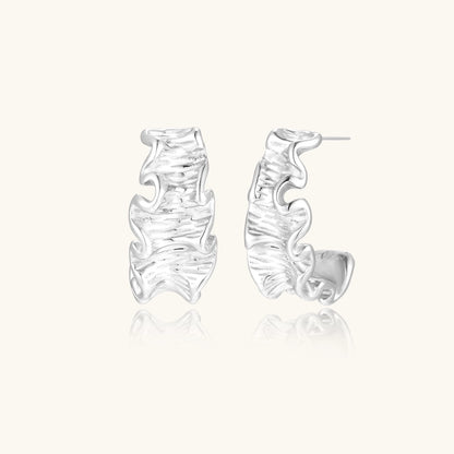 White gold plated sculptural hoop earrings with organic wavy edges and reflective metallic finish on cream background